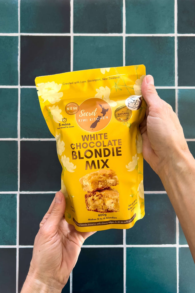 Hand holding a yellow package of white chocolate blondie mix against a jade tiled wall background